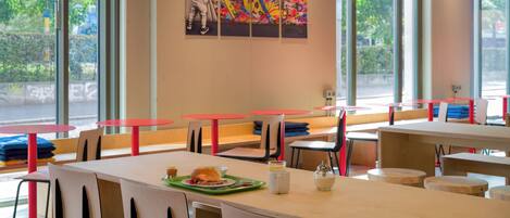 A dining area with tables and chairs, artwork on the wall, and large windows.
