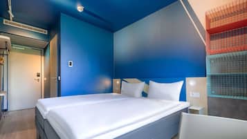 A modern hotel room with a large bed, a blue accent wall, and a built-in shelf with storage baskets.