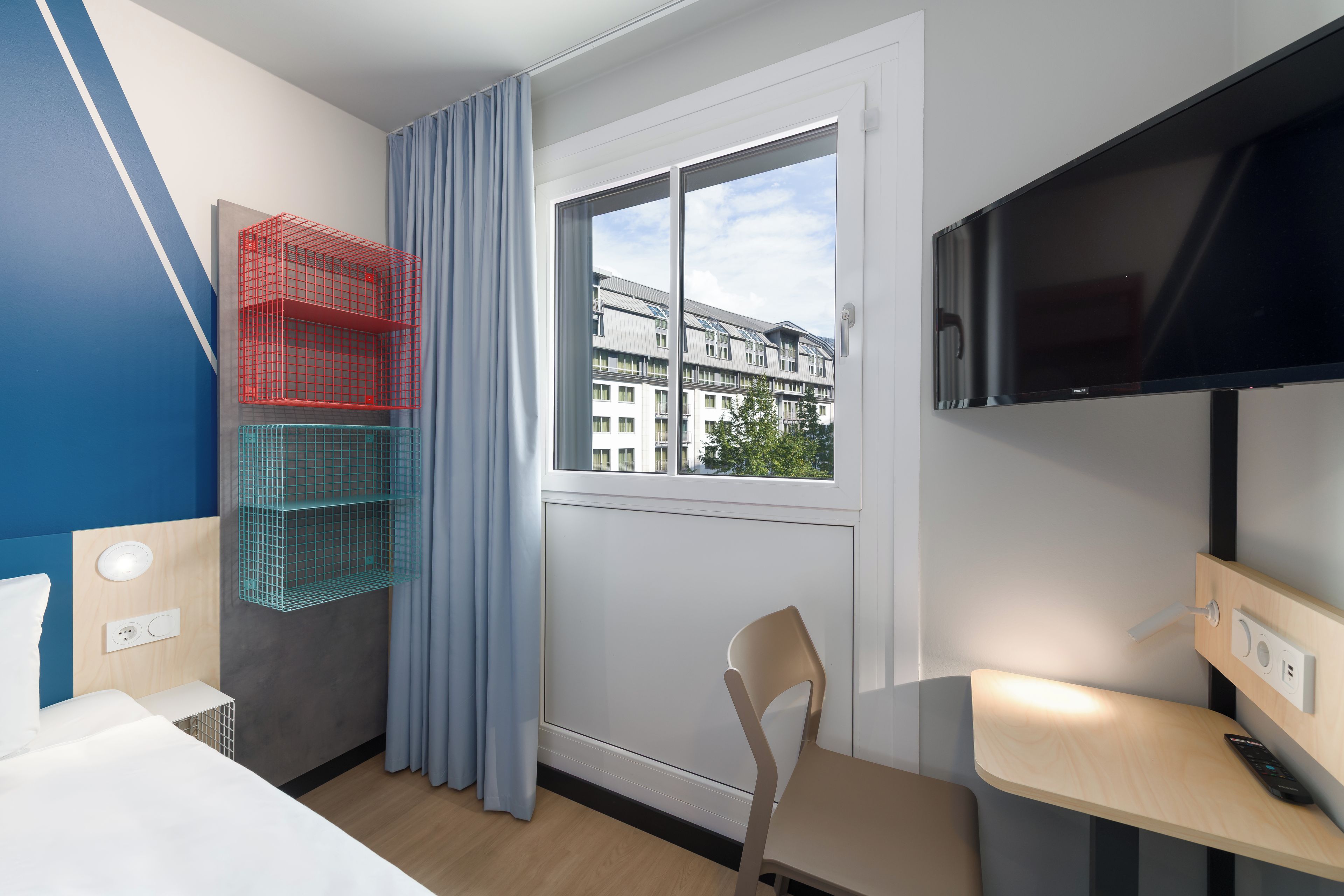 A hotel room with a bed, a desk with a chair, a television, and a window with a view of buildings.