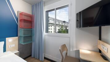 A hotel room with a bed, a desk with a chair, a television, and a window with a view of buildings.