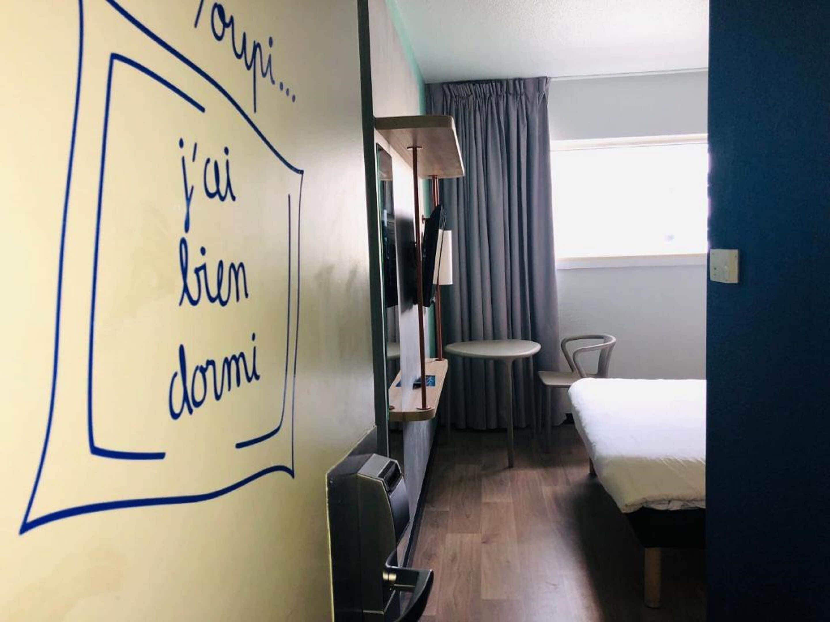 Une chambre d’hôtel avec un lit, un bureau, une chaise, une petite table et une lampe. Il y a une fenêtre avec des rideaux et un mur orné d’un dessin.