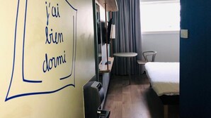 A hotel room with a bed, a desk, a chair, a small table, and a lamp. There is a window with curtains and a wall with a drawing. - ibis budget Paris Porte de Montreuil (Montreuil)