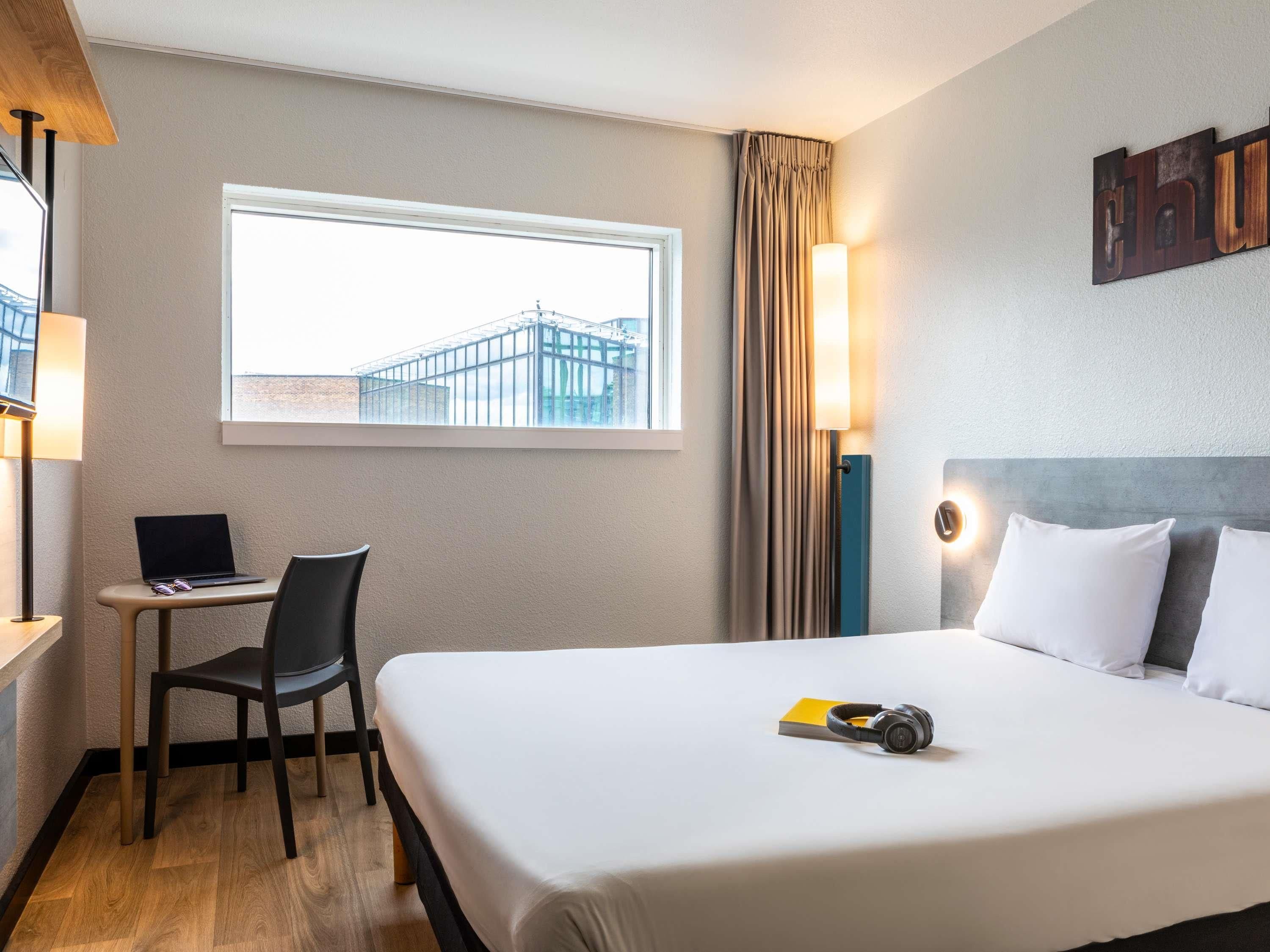 A hotel room with a bed, a desk with a laptop, a chair, a window with a view of buildings, and a wall-mounted light.