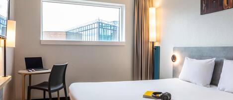 A hotel room with a bed, a desk with a laptop, a chair, a window with a view of buildings, and a wall-mounted light.
