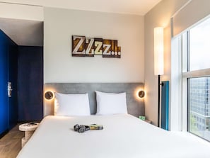 Hotelkamer met een bed, twee kussens, een nachtkastje met een koptelefoon en een boek, een wandbord en een raam met uitzicht op gebouwen.