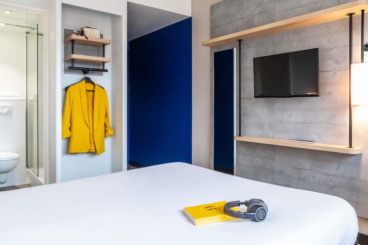A modern hotel room with a bed, a TV mounted on a grey wall, a yellow book, and headphones on the bed.