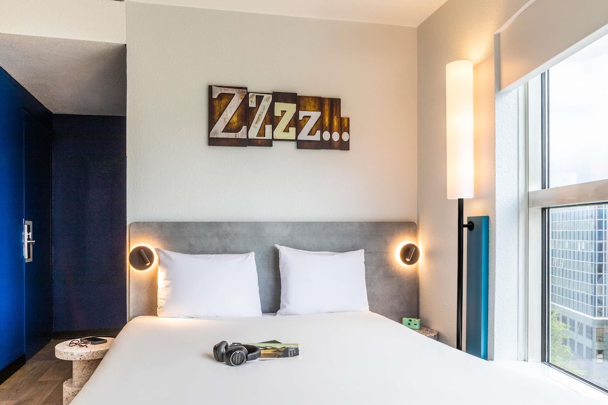 A hotel room with a bed, two pillows, a bedside table with a pair of headphones and a book, a wall-mounted sign, and a window with a view of buildings.