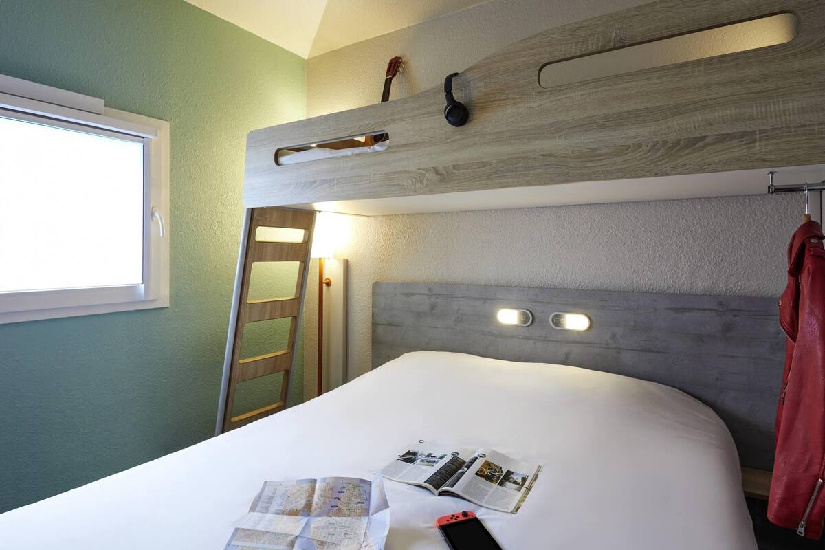 A bedroom with a bed, a window, a ladder, and a wall-mounted shelf with a guitar.