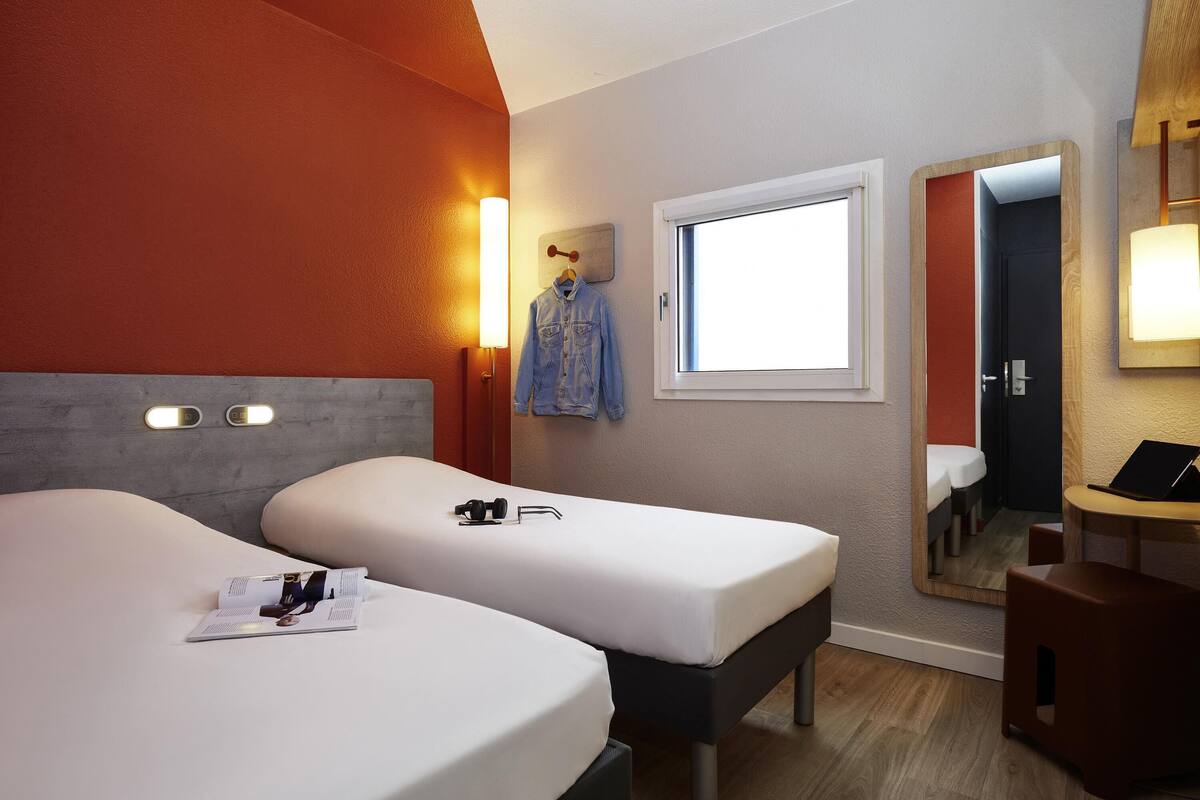 A hotel room with two beds, a window, a mirror, and a small desk with a laptop.