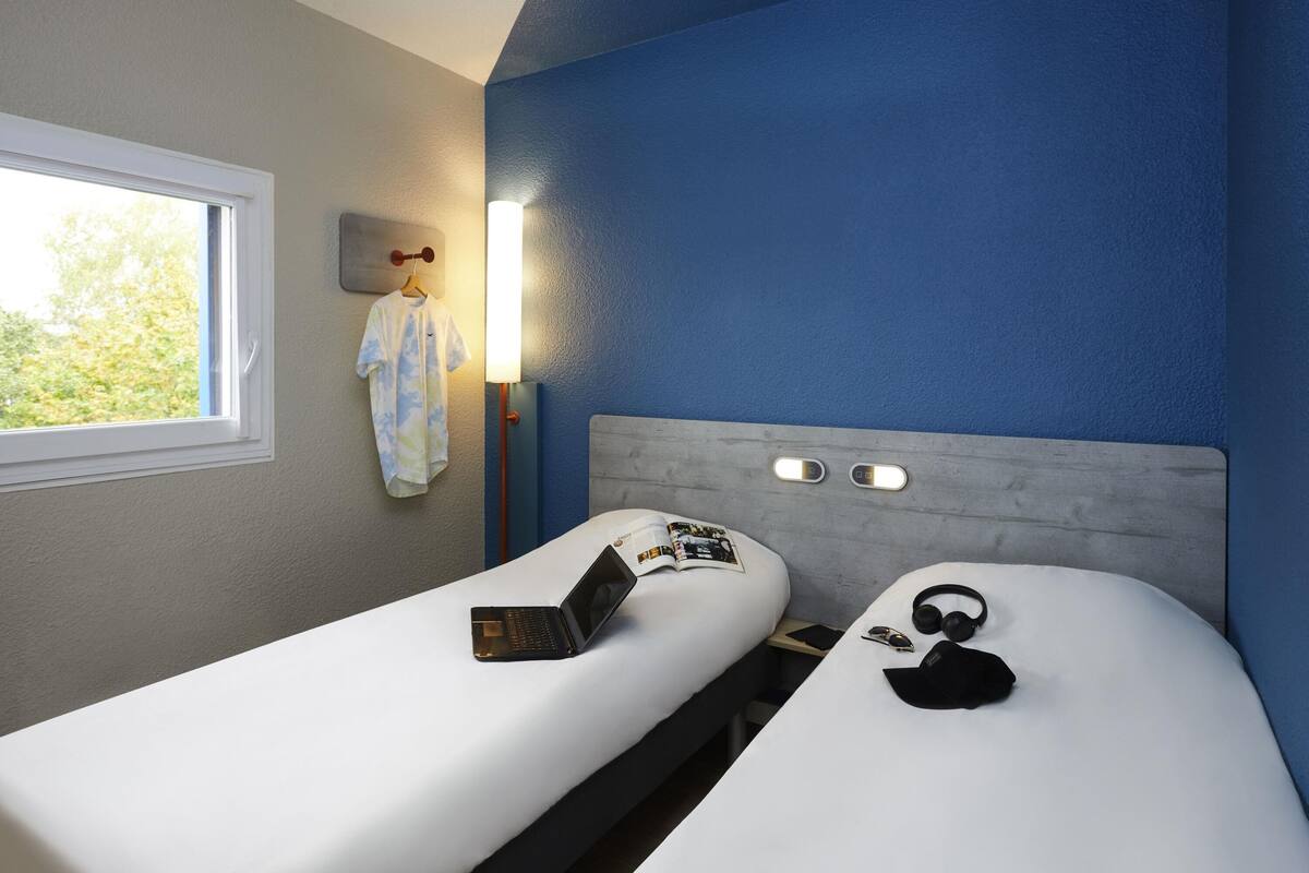 Habitación de hotel con dos camas individuales, una ventana, una lámpara fijada a la pared y una mesita con un ordenador portátil y un libro.