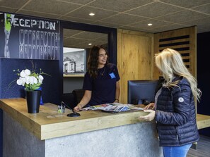 A reception desk with a sign reading 'RECEPTION' and a framed picture of a building. There is a potted plant on the counter. - ibis budget Lille Villeneuve d'Ascq (Villeneuve-d'Ascq)