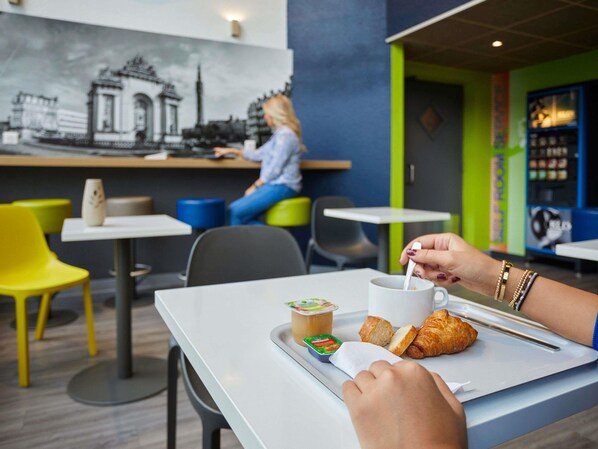 A café with a mural, tables, chairs, and a person eating. - ibis budget Lille Villeneuve d'Ascq (Villeneuve-d'Ascq)