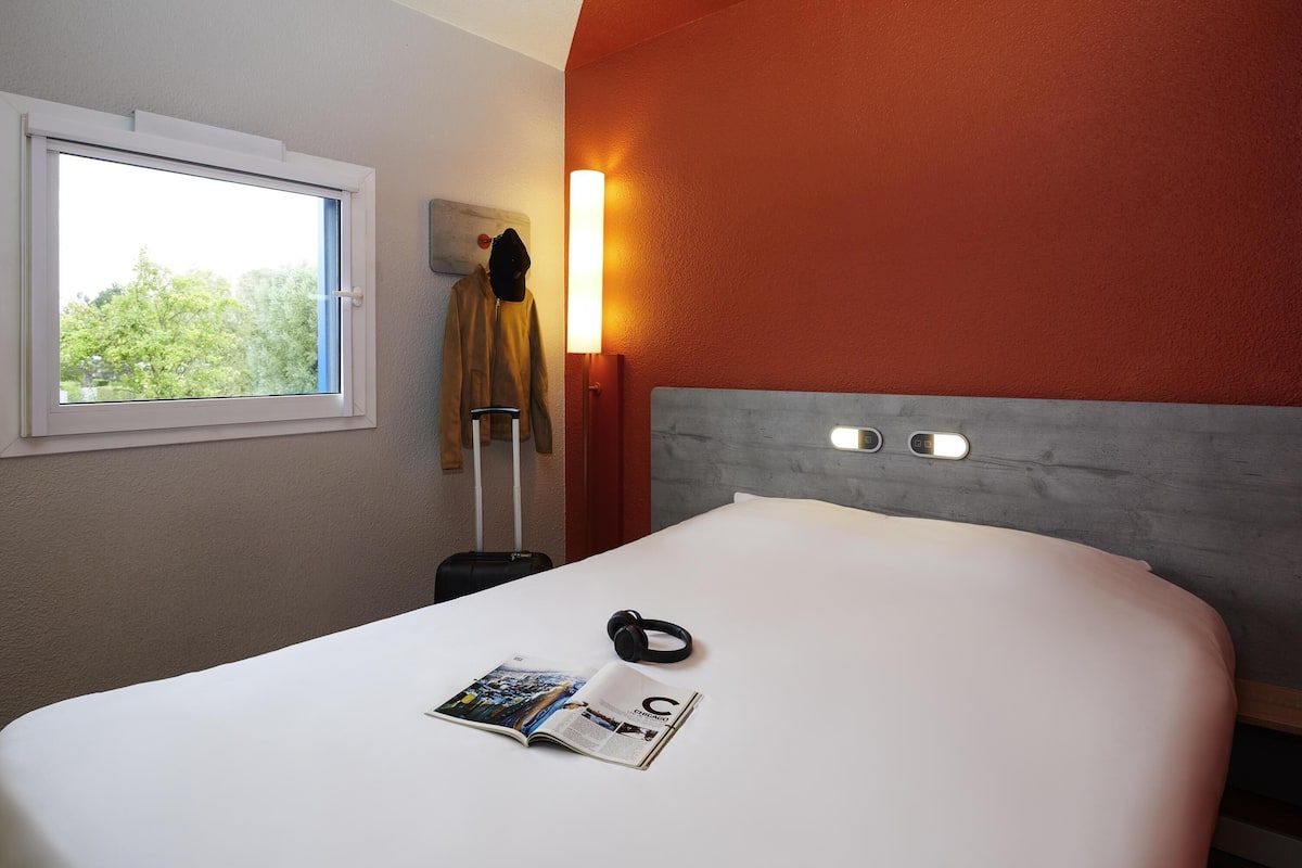 A hotel room with a bed, a window, a wall-mounted lamp, a coat rack, and a magazine on the bed.