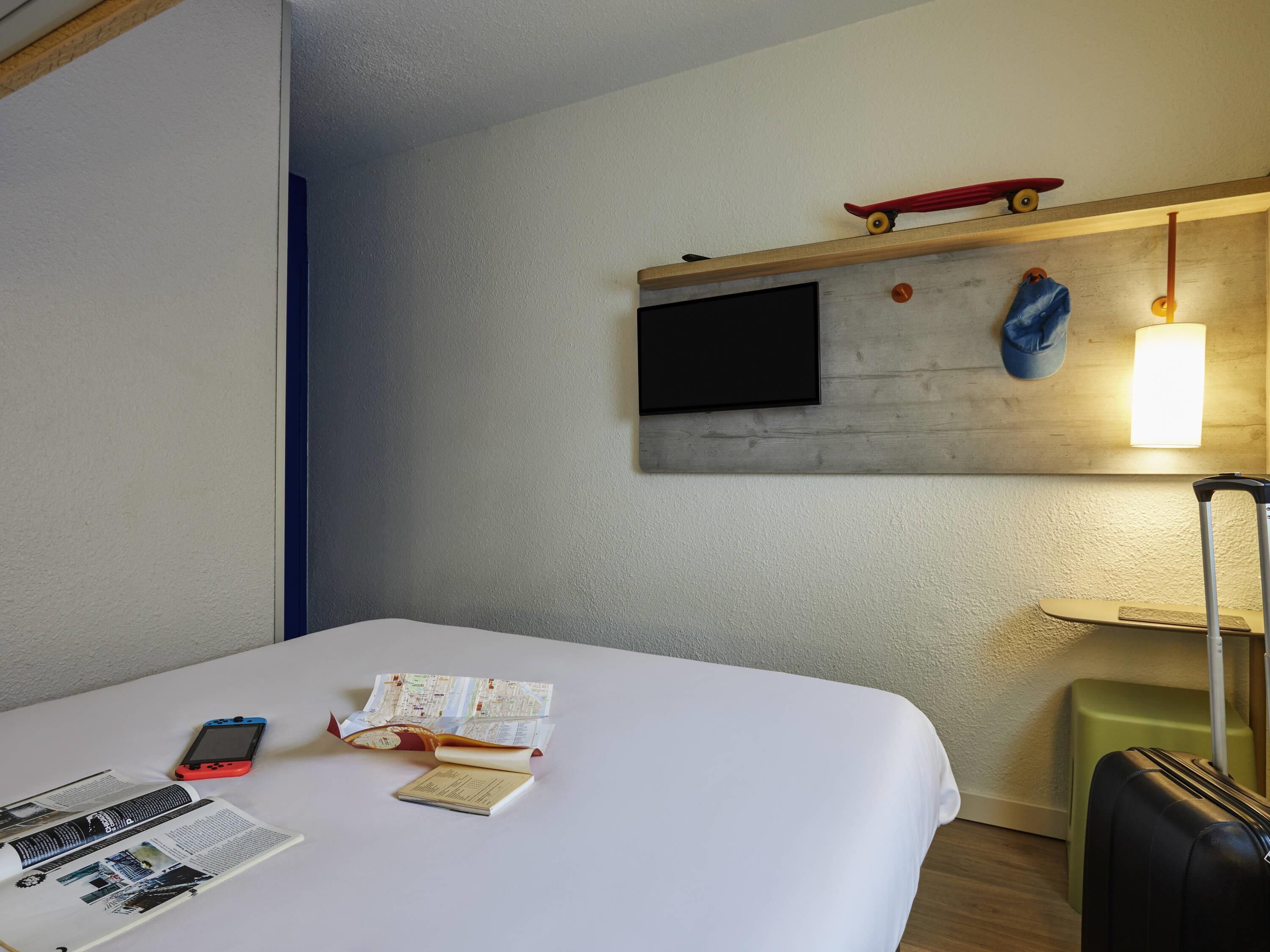Quarto de hotel com uma cama, televisão de tela plana na parede, um skate, um boné azul, uma mala e um abajur.