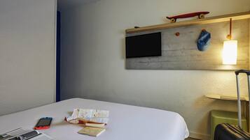 Quarto de hotel com uma cama, televisão de tela plana na parede, um skate, um boné azul, uma mala e um abajur.