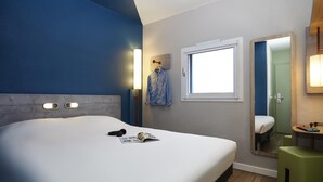 A hotel room with a bed, a window, a mirror, a bedside table, and a green stool. - ibis budget Lille Villeneuve d'Ascq (Villeneuve-d'Ascq)