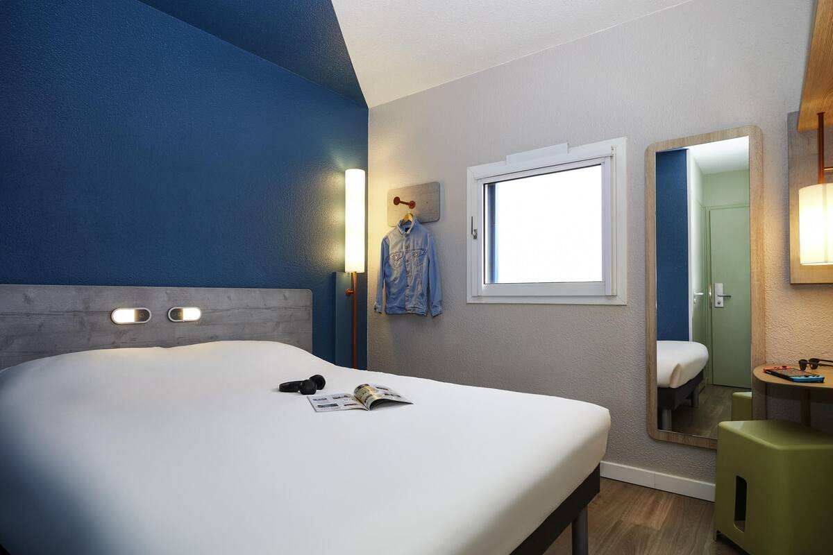 A hotel room with a bed, a window, a mirror, a bedside table, and a green stool.