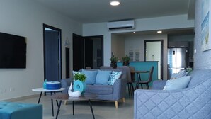 Una sala moderna con un sofá gris, un otomano azul y una mesa de centro. Se ve un área de cocina al fondo, con un refrigerador y un lavamanos.