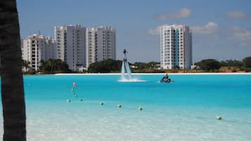 Una playa con agua turquesa cristalina, una fuente y edificios altos al fondo.