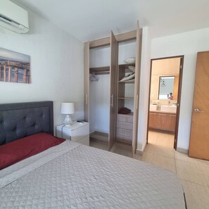 Un dormitorio con cama, mesita de noche, lámpara, armario y baño visible a través de una puerta abierta.