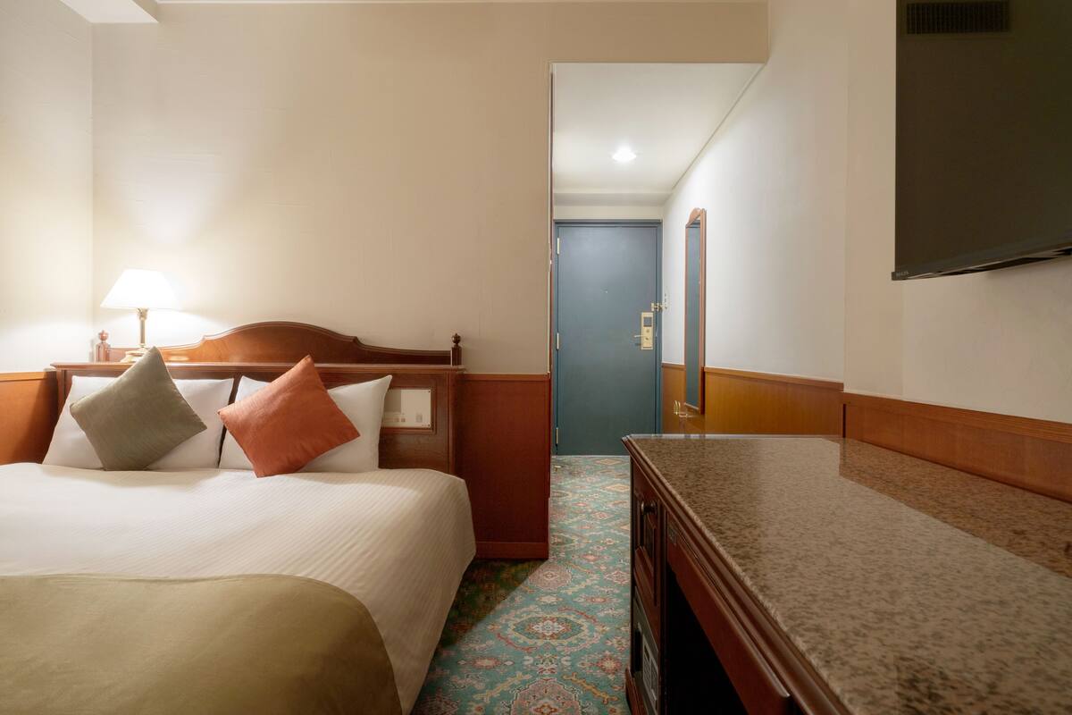 Habitación de hotel con cama, mesita de noche, lámpara, cómoda y televisor en la pared.