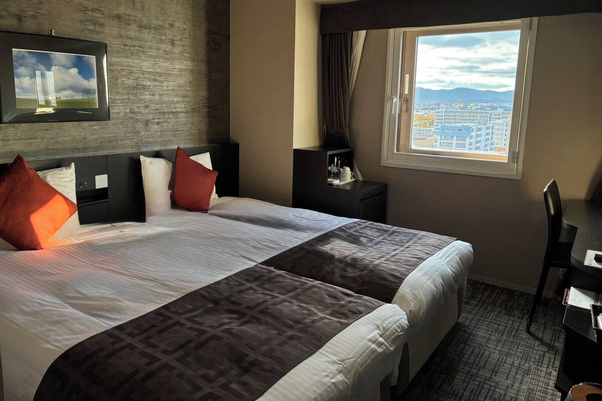 Habitación de hotel con una cama grande, un escritorio, una silla, un televisor montado en la pared y una ventana con vistas a la ciudad.