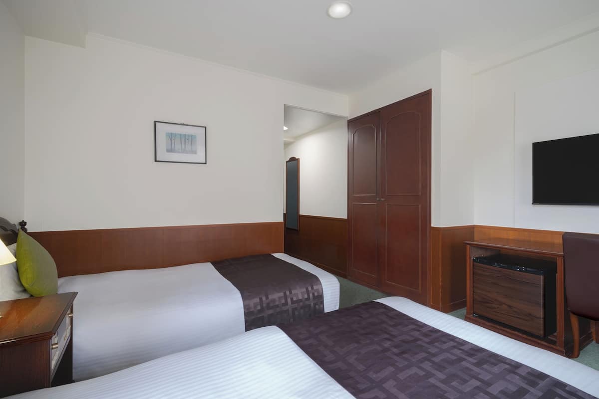 Habitación de hotel con dos camas, un armario de madera, un televisor montado en la pared y un cuadro enmarcado.