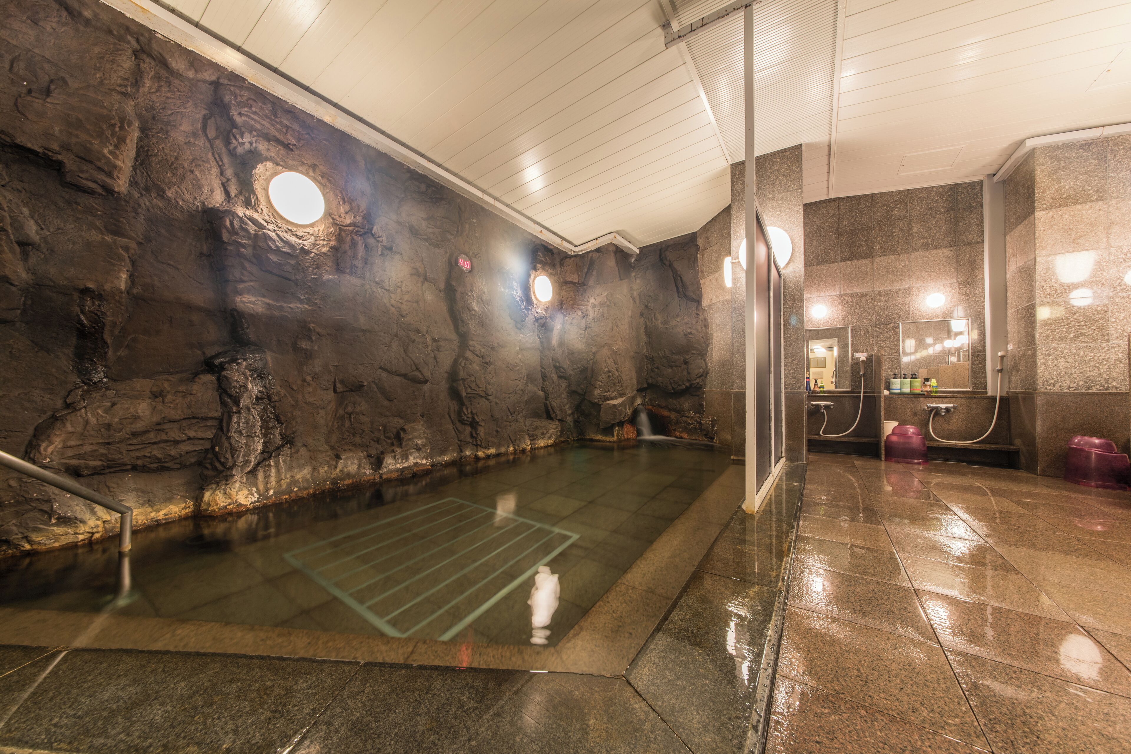 A stone-walled hot spring bath with a glass floor, a shower area, and a mirror.