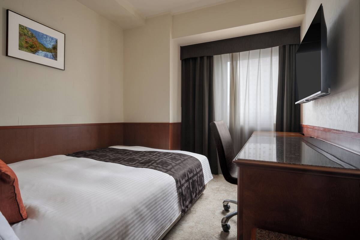 Una habitación de hotel con cama, escritorio, silla, televisor y ventana con cortinas.