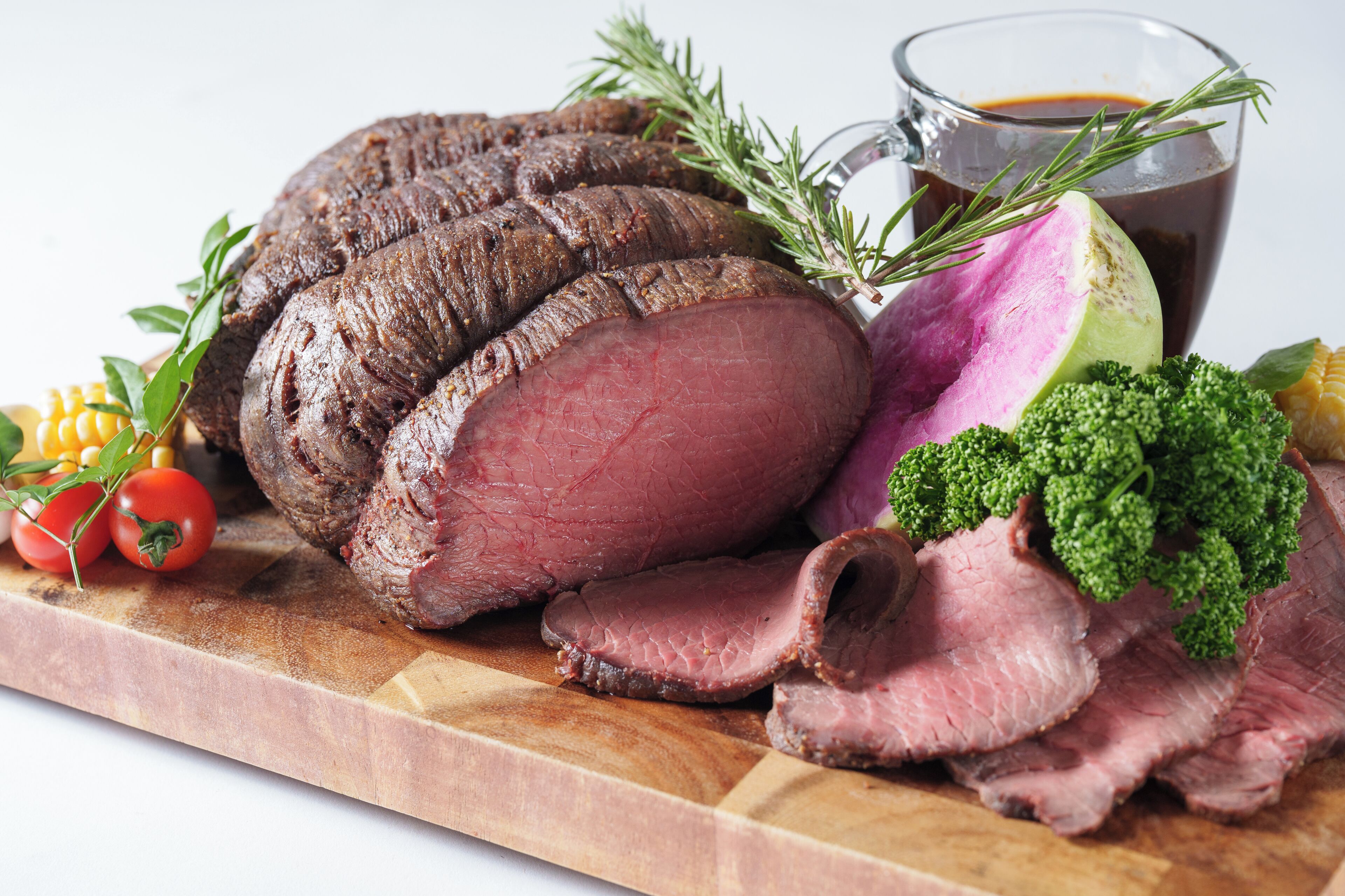 A wooden cutting board with sliced roast beef, fresh vegetables, and a glass of dark liquid.