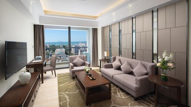 A modern hotel room with a large window, a flat-screen TV, a sofa, a coffee table, and a small side table with a vase of flowers. - Sanya Shuangda Adelaide In Resort Hotel (Sanya)