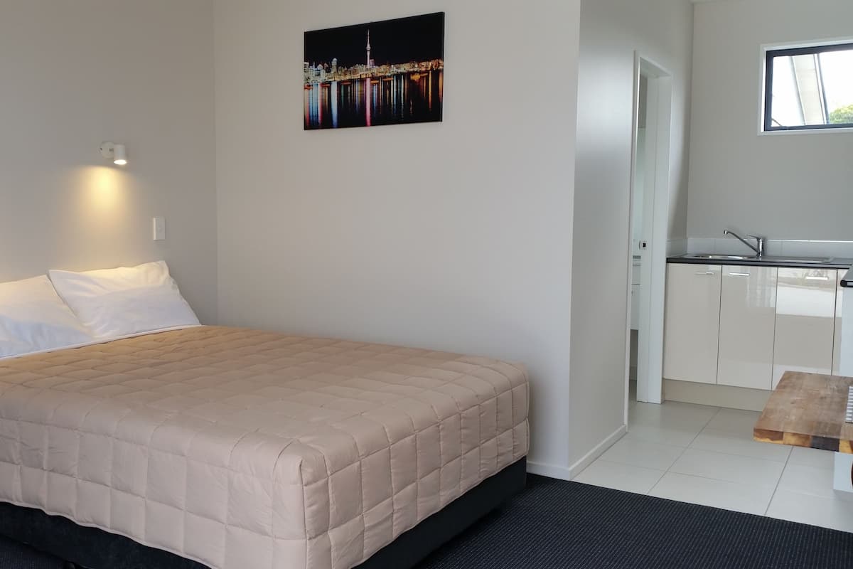 A hotel room with a bed, two bedside lamps, a nightstand, a small table, a picture on the wall, and a kitchenette with a sink and cabinets.