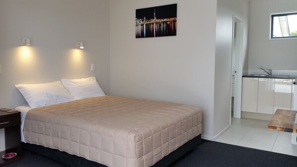 Quarto de hotel com cama, duas luminárias de cabeceira, mesa de cabeceira, uma pequena mesa, um quadro na parede e uma cozinha compacta com pia e armários.