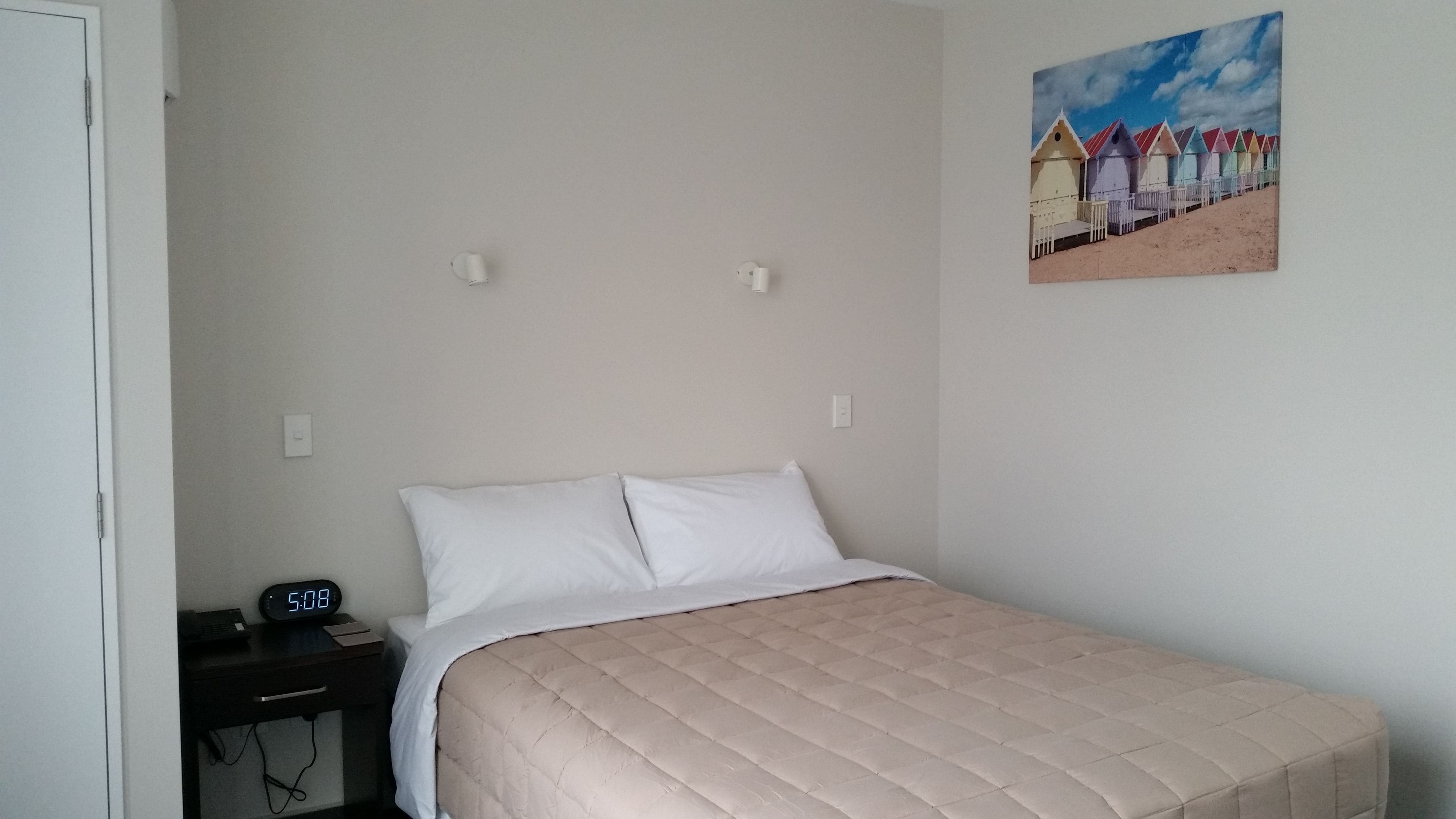 Une chambre à coucher avec un lit, une table de chevet sur laquelle se trouve une horloge, et un tableau représentant des cabanes de plage colorées accroché au mur.