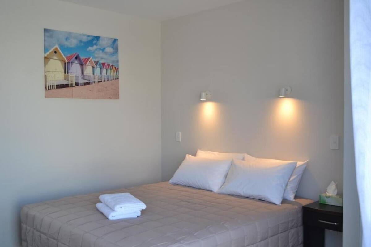 A bedroom with a bed, pillows, a nightstand, a painting, and wall-mounted lights.