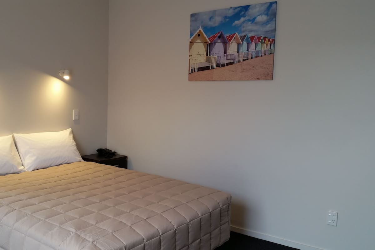 A neatly made bed with a beige quilt, a nightstand with a phone, and a painting of colorful beach huts on the wall.