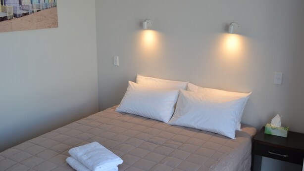 A neatly made bed with white bedding and two pillows, a bedside table with a tissue box, and a painting of beach huts on the wall. - 319 Addington Motel (Christchurch)