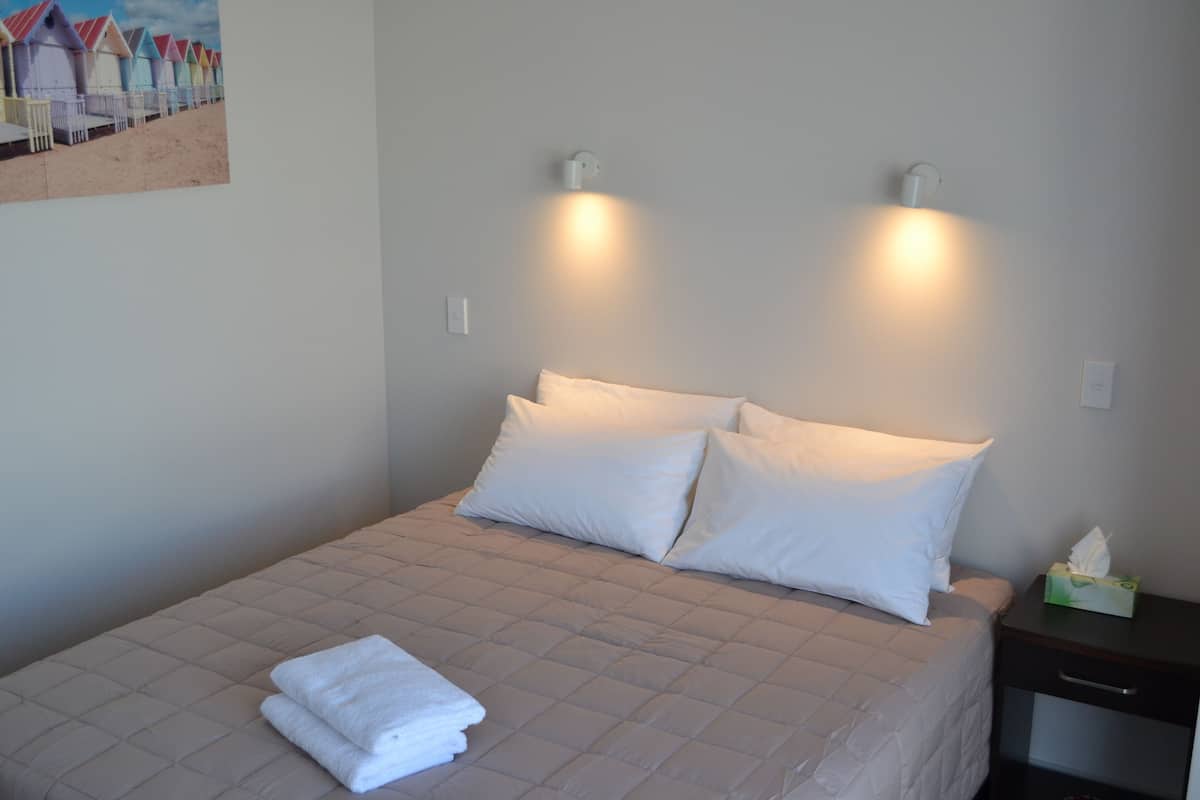 A neatly made bed with white bedding and two pillows, a bedside table with a tissue box, and a painting of beach huts on the wall.