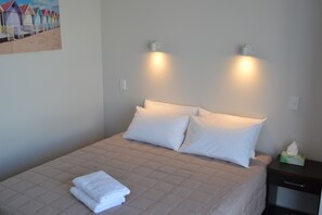 A neatly made bed with white bedding and two pillows, a bedside table with a tissue box, and a painting of beach huts on the wall.