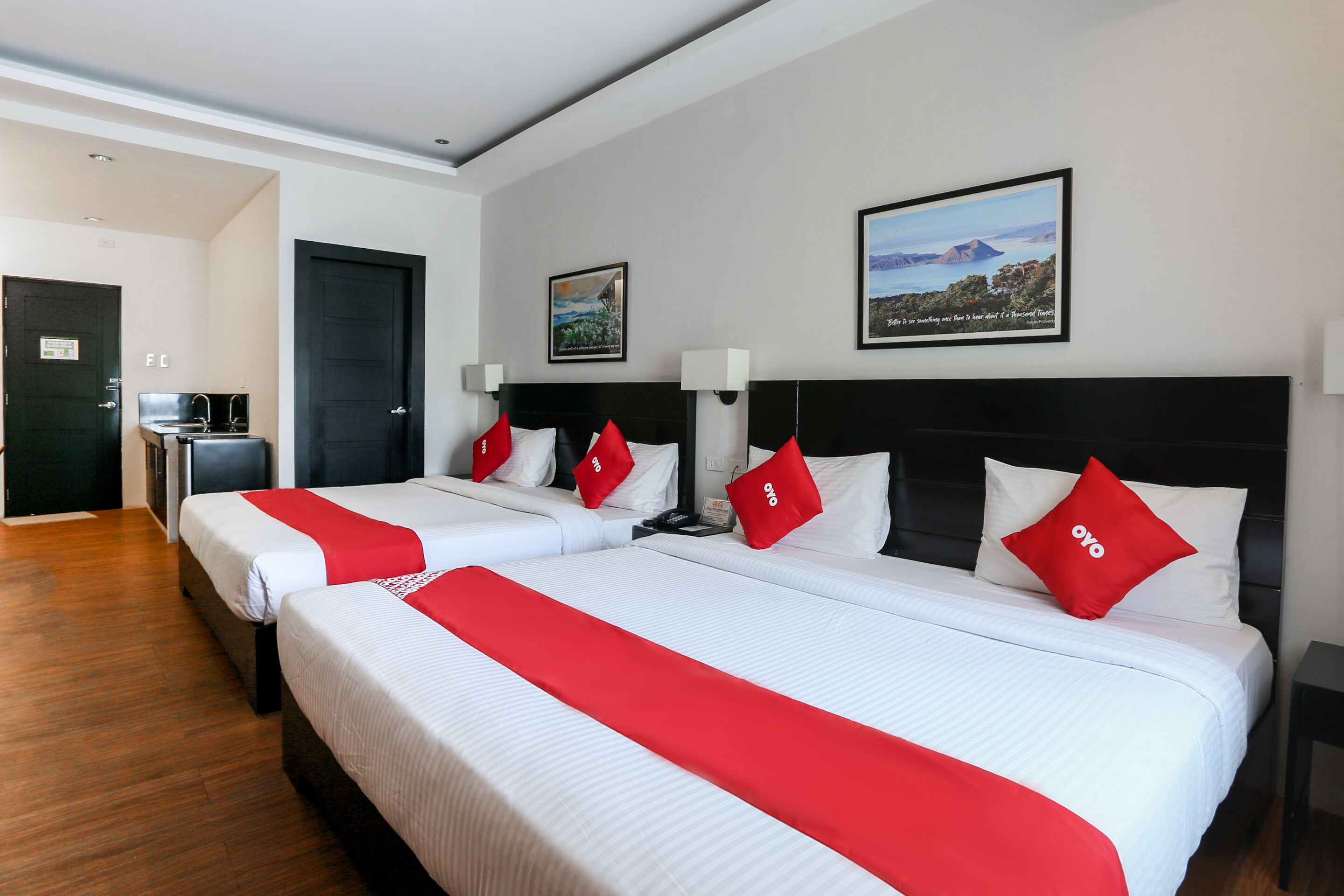 A hotel room with two beds, each with white bedding and red accents, a black headboard, a framed landscape picture on the wall, and a wooden floor.