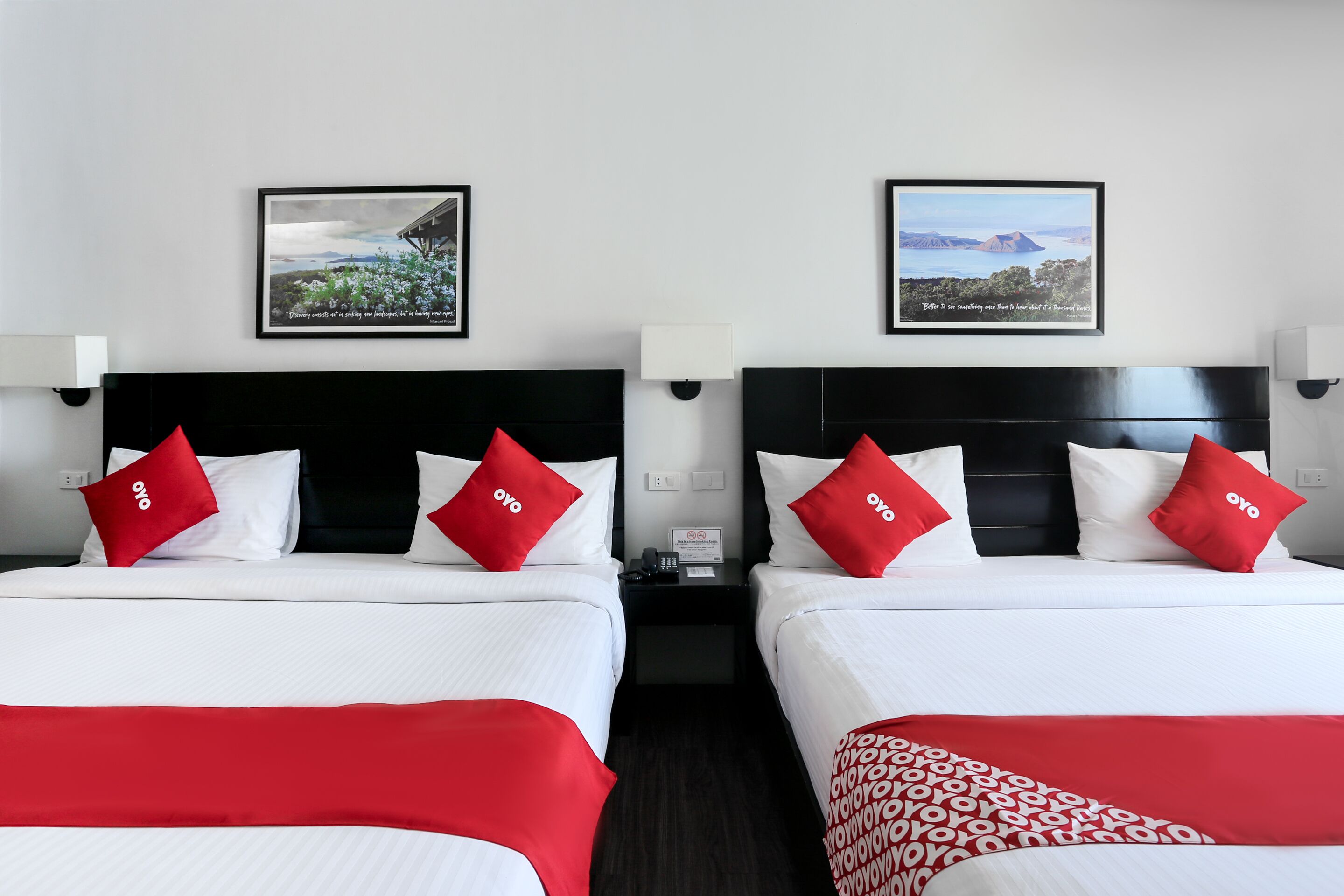Habitación de hotel con dos camas, cada una con ropa blanca y almohadas rojas, y una colcha roja. Encima de las camas, hay cuadros enmarcados en la pared.