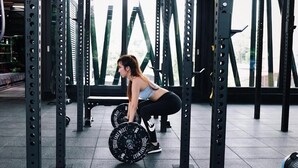 Una mujer realiza un ejercicio de sentadilla con barra en un gimnasio equipado para levantamiento de pesas y con amplios ventanales.