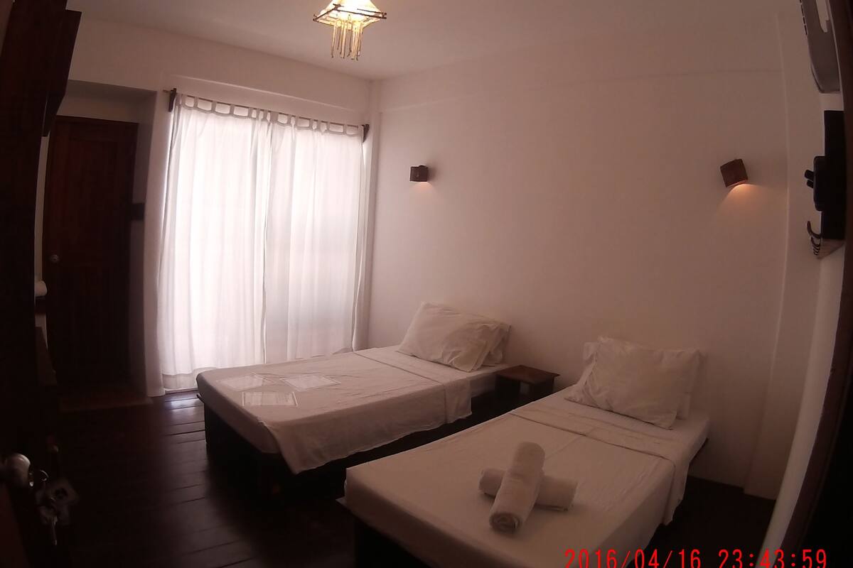 Habitación de hotel con dos camas individuales, piso de madera, ventana con cortinas y una lámpara colgante.