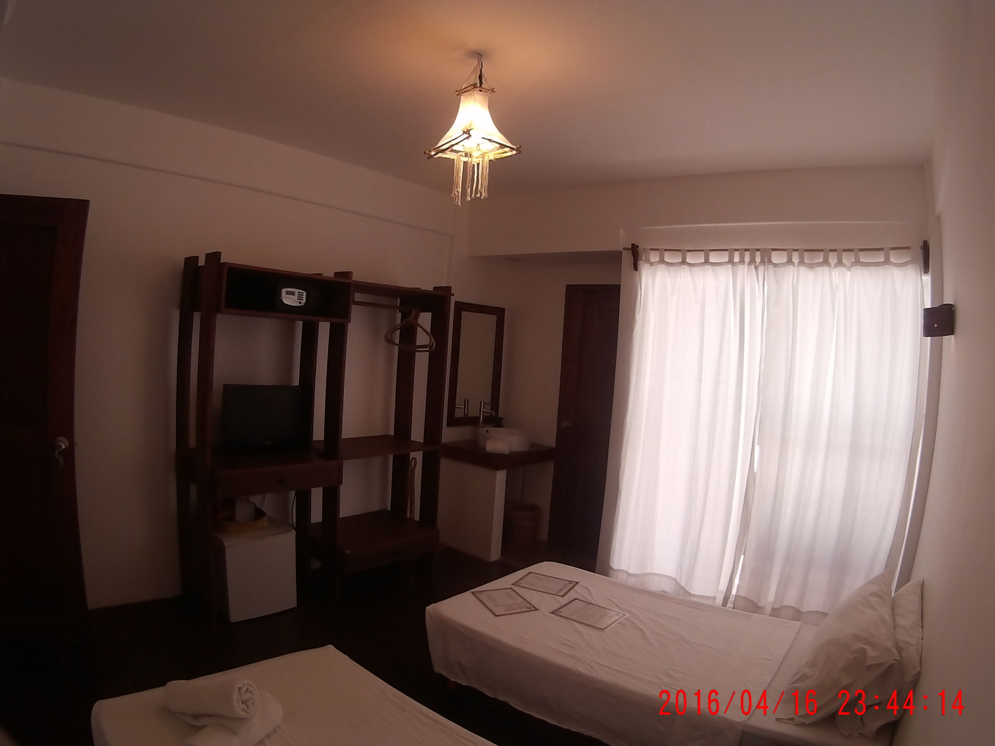 A hotel room with a bed, a bedside table, a mirror, a hanging light fixture, and a window with curtains.