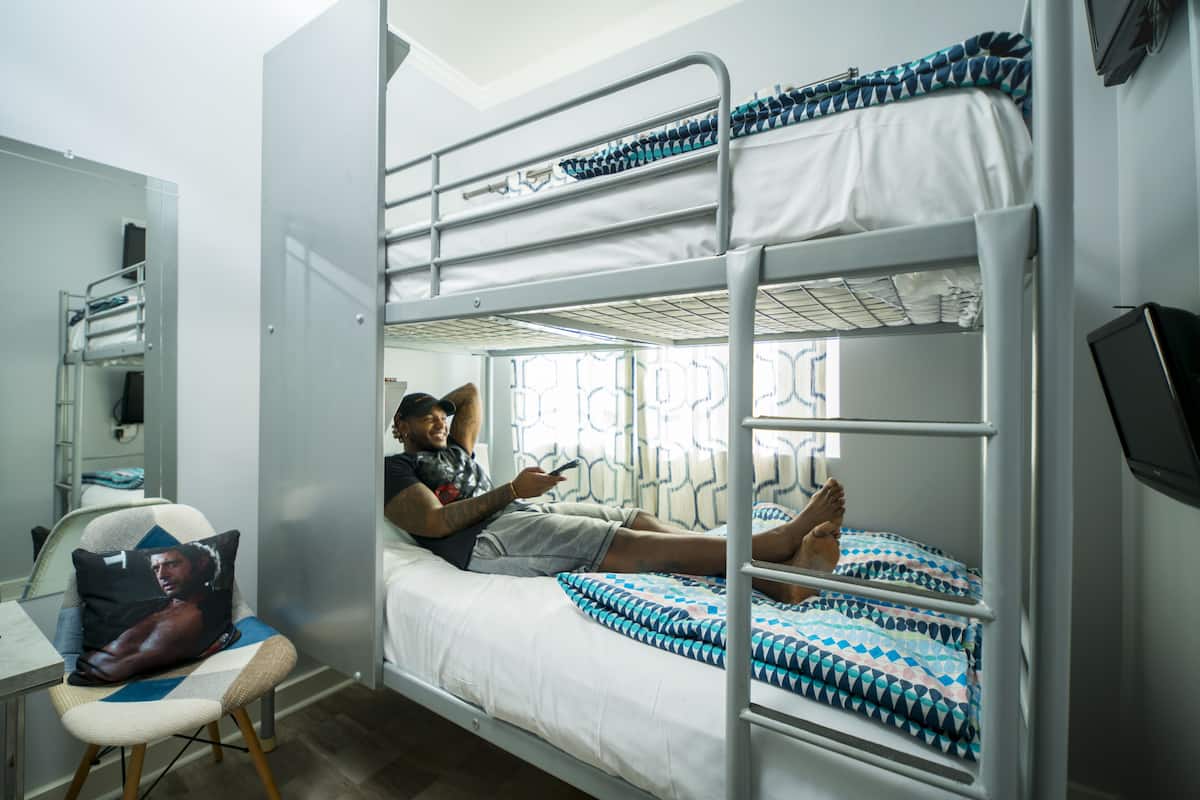 Quarto com beliche, uma pessoa deitada no andar superior, uma cadeira com almofada e janela com cortinas estampadas.