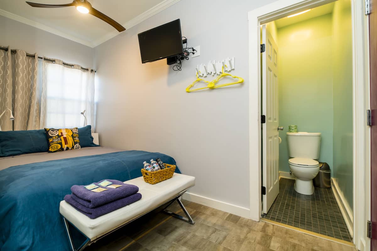 Quarto de hotel com uma cama, um banco com toalhas, uma televisão fixada na parede e um banheiro com vaso sanitário e chuveiro.