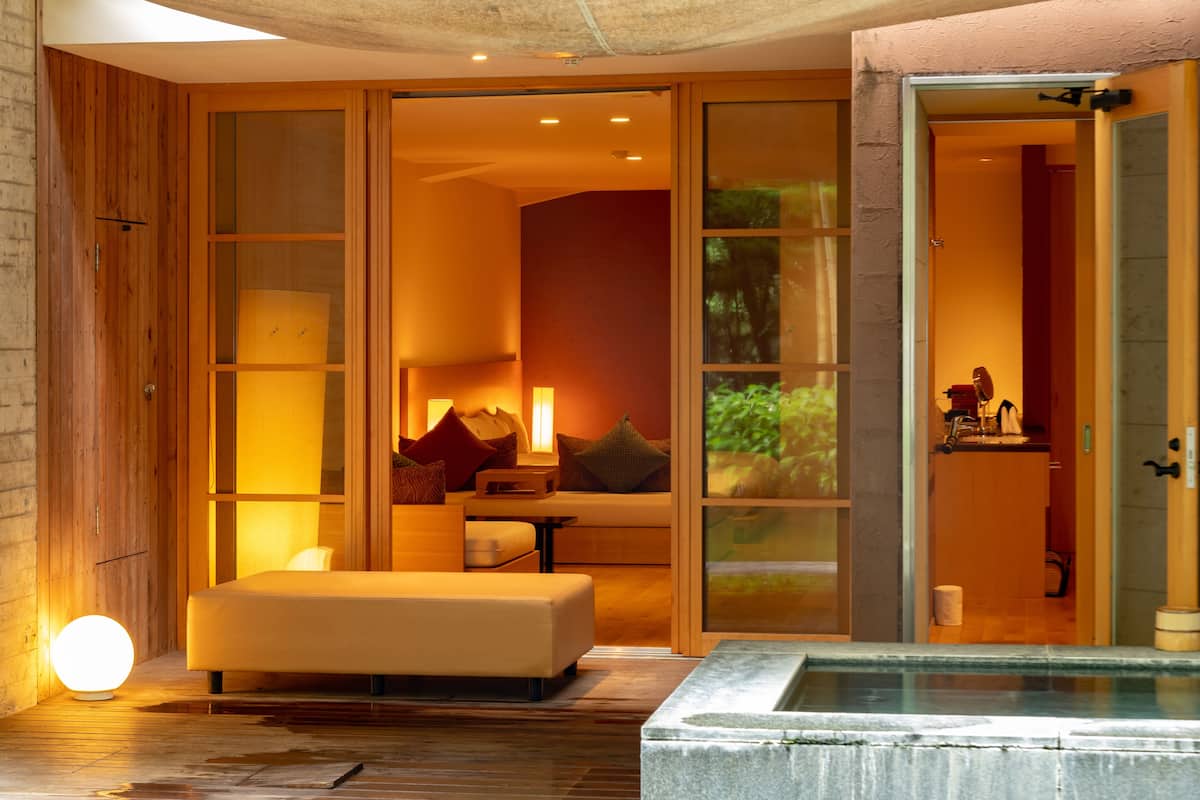 A modern hotel room with a large sliding glass door leading to a living area, a cozy seating area with cushions, and a visible bathroom.