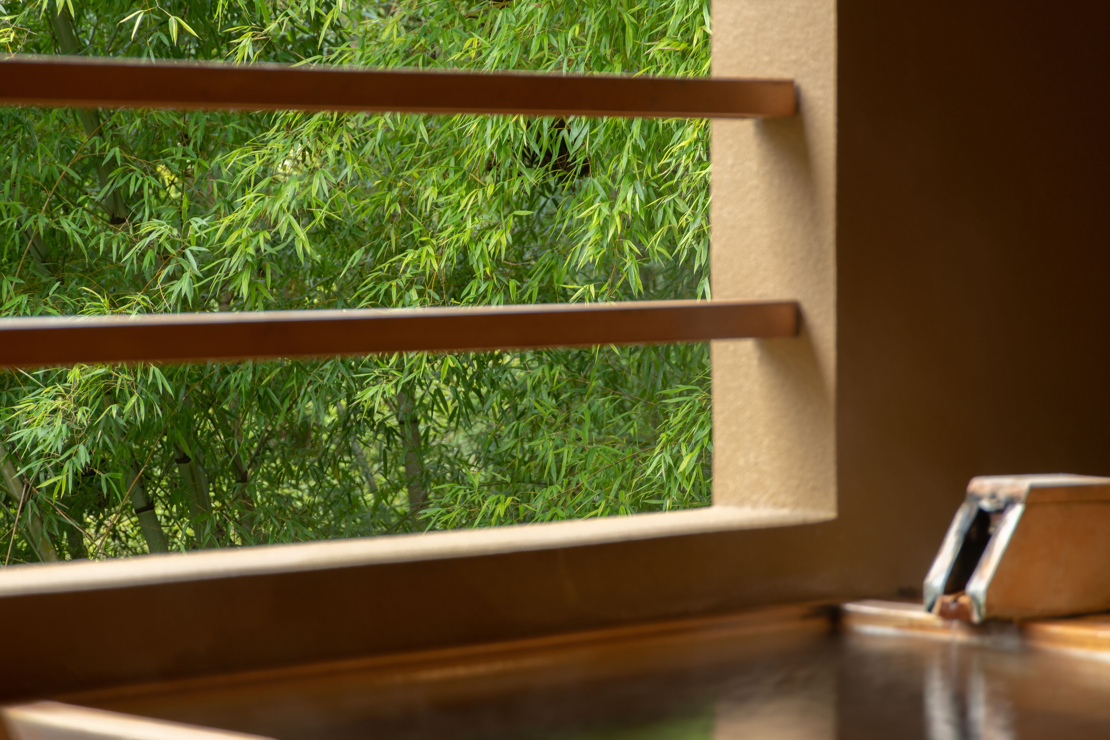 Una ventana con persianas horizontales, una mesa de madera y vistas a follaje verde.