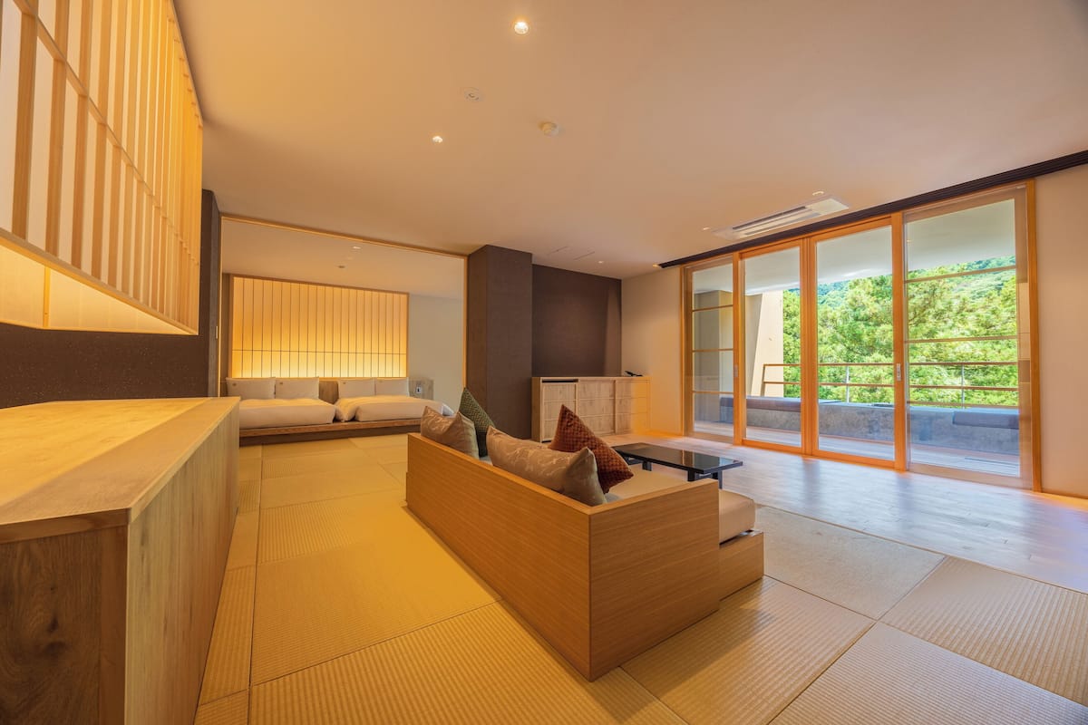 A modern interior with a wooden reception desk, a sofa with cushions, a cabinet, and a large sliding glass door leading to a balcony with a view of greenery.