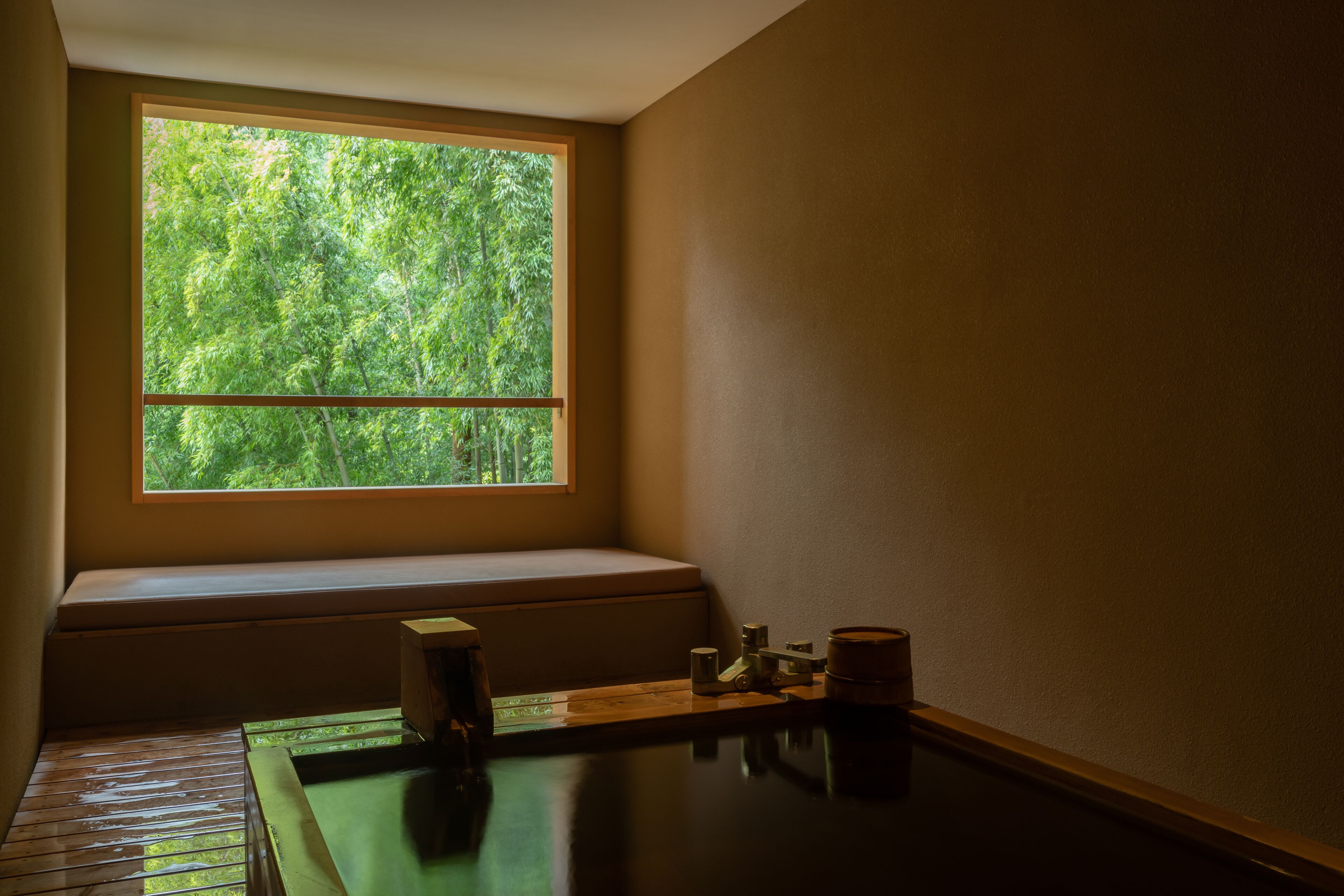 A room with a large window overlooking a forest, a wooden bench, and a small table with a teapot and cups.